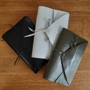 Three leather notebooks in white, green, and black leather. They all have a triangular cover flap and ties that wrap round and hold it shut.