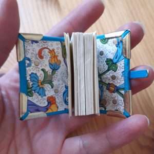 A picture to advertise a miniature bookbinding class. A hand holding a miniature book with metal corners and a clasp, and brightly coloured endpapers.